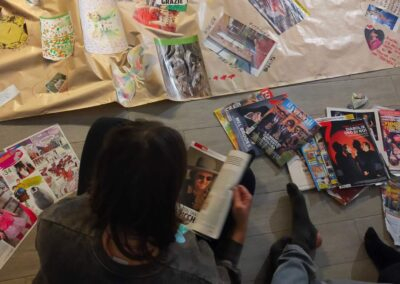 Vista dall'alto di un lungo cartellone con collage, farfalle disegnate e parole come "grazie", accanto a riviste