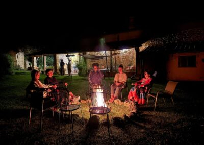 Gruppo di donne sedute in cerchio attorno a un fuoco acceso di sera nel giardino di un casale durante un ritiro