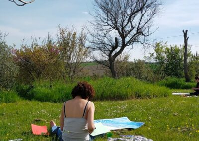Donna di spalle seduta in un prato fiorito dipinge su grandi fogli in una giornata di sole primaverile