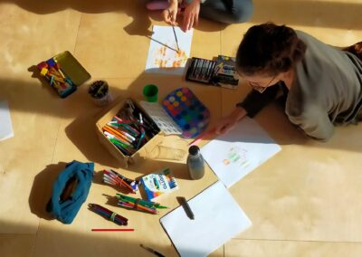 Due donne disegnano e dipingono sedute a terra, immerse nella luce naturale, durante un laboratorio espressivo