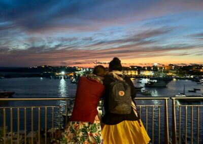 Due donne di spalle affacciate a una ringhiera guardano il tramonto sul porto di Lampedusa illuminato dalle barche