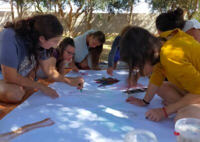 Gruppo di donne disegna insieme su un grande foglio steso a terra sotto gli ulivi durante un laboratorio all'aperto