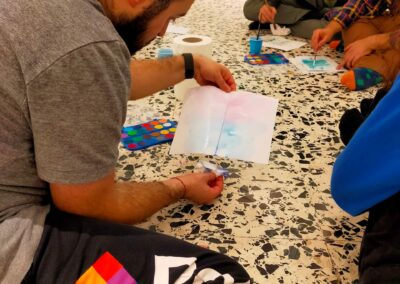 Partecipante osserva un acquerello appena dipinto mentre un gruppo dipinge seduto a terra in un laboratorio