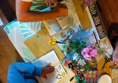Vista dall'alto di un tavolo con libri, scrittura a mano, pastelli, fiori e mani al lavoro in un laboratorio