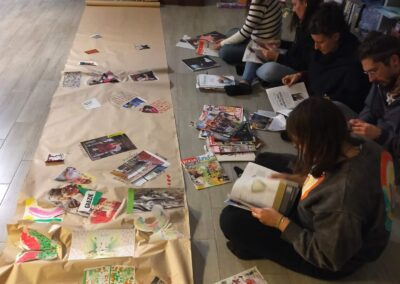 Gruppo seduto a terra sfoglia riviste accanto a un lungo cartellone con collage e disegni in una biblioteca